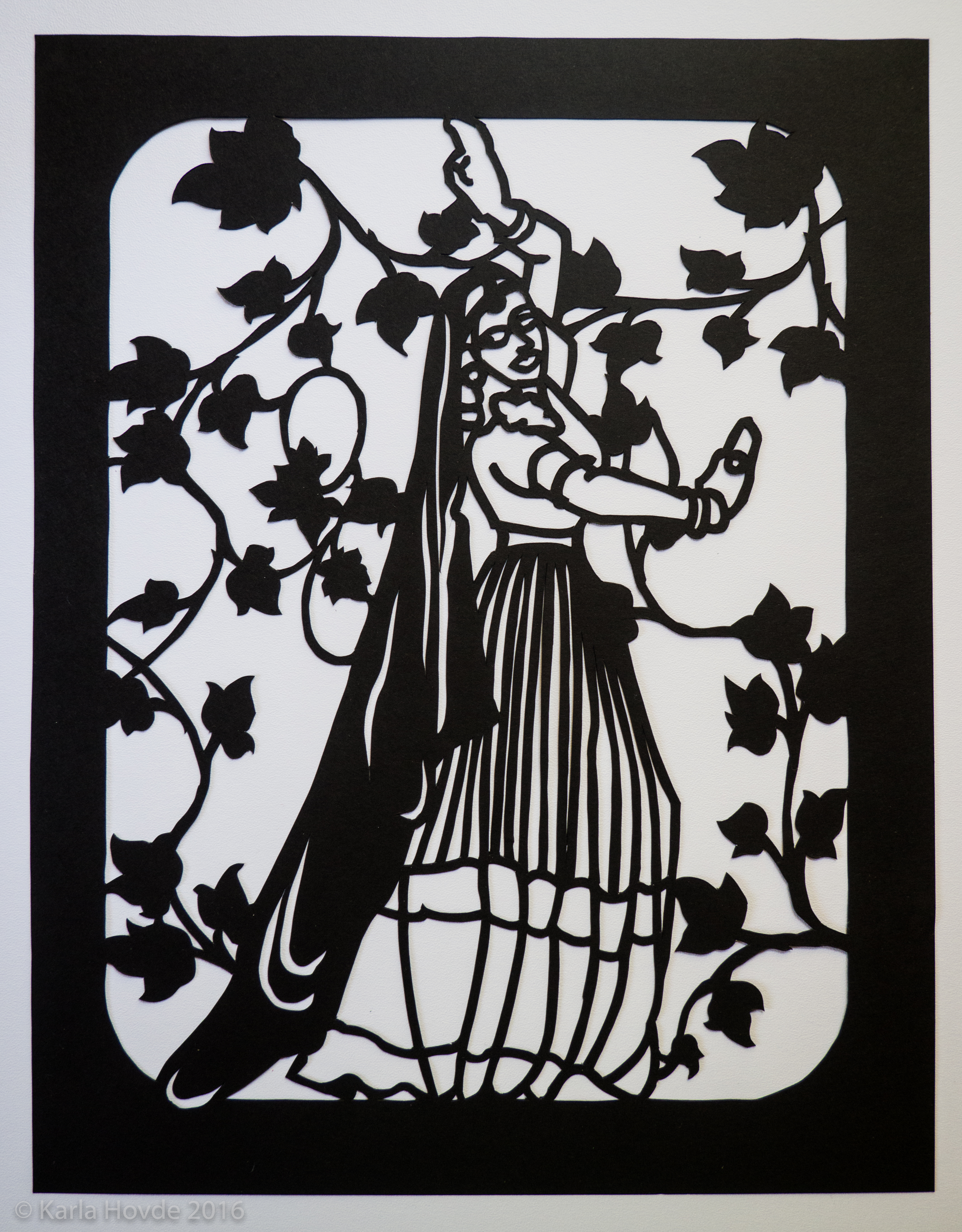 Paper cutting art in black paper shows stylized woman wearing a sari and dancing in front of delicate ivy leaves pattern.